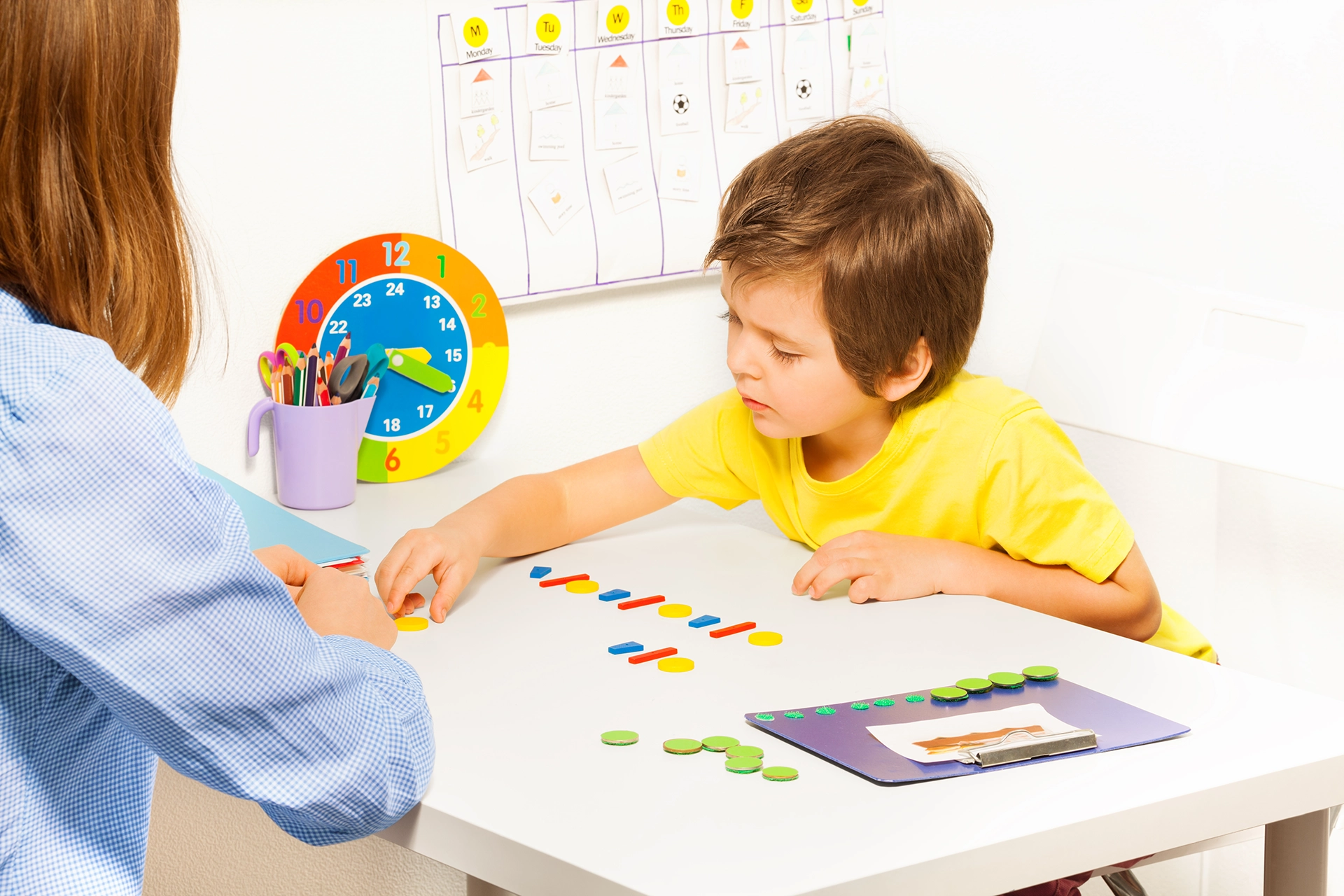 Concentrated boy putting colorful coins in order during developing game with a teacher sitting at the table